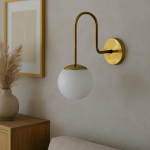 May include: A contemporary wall light featuring a gold-toned metal frame and a spherical, frosted glass shade. The fixture is mounted on a wall, with a curved arm and a circular backplate. The design is minimalist, suitable for interior lighting.
