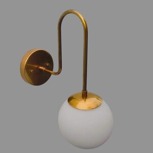 May include: A gold-coloured wall sconce featuring a curved arm and a white globe shade. The sconce has a circular base and a decorative design. The light fixture is a modern design and is suitable for indoor use.