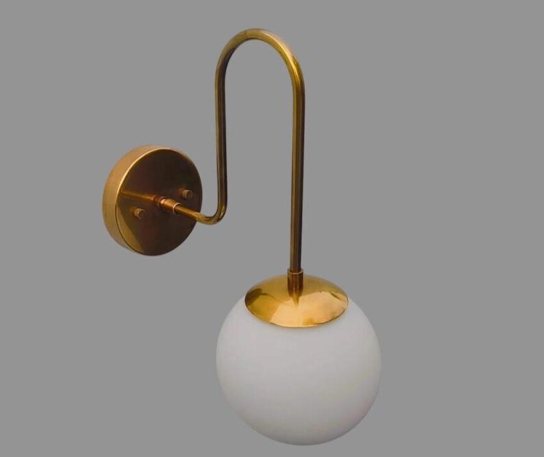 May include: A gold-coloured wall sconce featuring a curved arm and a white globe shade. The sconce has a circular base and a decorative design. The light fixture is a modern design and is suitable for indoor use.