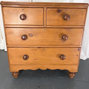 May include: A light brown wooden chest of drawers featuring two small top drawers and two larger bottom drawers. Each drawer is equipped with a round wooden knob and a central keyhole. The piece stands on four turned bun feet.