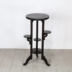 Może przedstawiać: Ciemnobrązowy drewniany stojak na rośliny z okrągłym blatem i trzema mniejszymi okrągłymi półkami. Stojak ma trzy nogi i dekoracyjny wzór na krawędziach półek. Stojak ma około 76 cm wysokości.