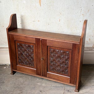 May include: A low wooden cabinet featuring two doors with intricate carved floral patterns in a diamond grid layout. The piece has a flat top surface, raised side panels, and a dark brown wood grain finish with a small metal keyhole in the centre.