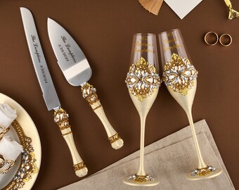 May include: A wedding cake knife and server set with matching champagne flutes. The knife and server have cream-colored handles with gold and crystal accents. The flutes are decorated with gold and crystal designs. The set includes two gold wedding bands.