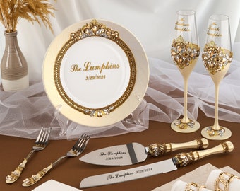 May include: A collection of wedding accessories, including a decorative plate, champagne flutes, cake server, and forks. The plate and flutes are adorned with gold and crystal embellishments. The plate reads "The Lumpkins 3/29/2024".