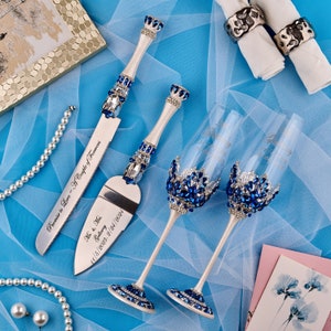 May include: A wedding cake cutting set with two champagne flutes and a knife. The flutes and knife are decorated with blue and silver rhinestones. The knife has the text "Promise to Love - A Couple of Forevers" and "Mr. & Mrs. Galloway 11/5/2023 - 2/24/2024" engraved on it.