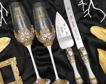 May include: A wedding cake cutting set with two champagne flutes and a cake knife. The flutes and knife are decorated with rhinestones and gold accents. The flutes are engraved with "Mr. & Mrs. Lakkis 05.21.23". The cake knife is engraved with "Mr. & Mrs. Lakkis 05.21.23".