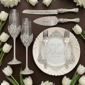 May include: Wedding table setting with champagne flutes, cake knife and server, plate, and forks. The items are decorated with rhinestones and the names "Mr & Mrs Khalid" and the date "04/16/2024" are engraved. White tulips and flowers surround the items.
