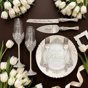 May include: A wedding table setting featuring engraved champagne flutes, cake server, and plate. The flutes and plate are decorated with sparkling details. The cake server and plate are engraved with "Mr. & Mrs. Khalil" and the date 16/04/2024. White tulips and a ribbon complete the arrangement.
