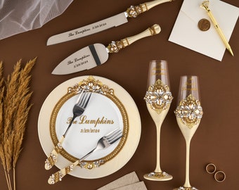 May include: A wedding cake cutting set with two gold-handled knives, two champagne flutes with gold and crystal embellishments, and a white plate with gold trim and the text "The Lumpkins 3/29/2024".