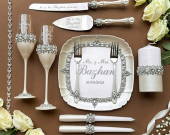 May include: Wedding cake cutting set with a white cake plate, two champagne flutes, two candles, and two cake knives. The cake plate and flutes are decorated with rhinestones. The cake knives are engraved with "Mr. & Mrs. Bazhan 10/09/2023".