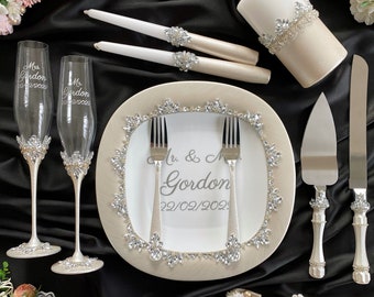 May include: Wedding decor set with two champagne flutes, a cake knife and server, two candles, and a plate with a white background and a silver trim. The flutes and plate are decorated with rhinestones. The plate has the text "Mr. & Mrs. Gordon 22/02/2022" written on it.
