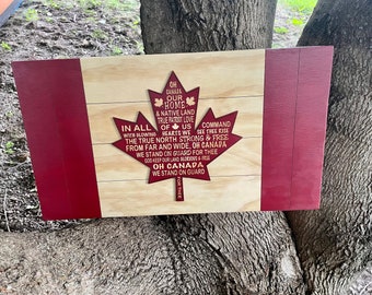 May include: Wooden Canadian flag with a red maple leaf and red borders. The maple leaf contains the lyrics of the Canadian national anthem. The flag is made of light-colored wood.