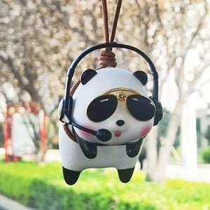 May include: A white panda bear wearing black headphones and sunglasses, with a brown cord hanging from its head. The panda is a car charm.