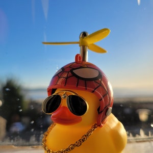 Puede incluir: Un pato de goma amarillo que lleva un casco rojo y negro con un diseño de telaraña, gafas de sol y una cadena de oro. El pato tiene una hélice amarilla en la cabeza.