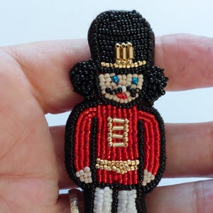 May include: A beaded ornament in the shape of a toy soldier. The soldier has a black hat and hair, a red jacket, and black pants. The soldier's face has blue eyes and a red mouth. Gold beads are used for the details.