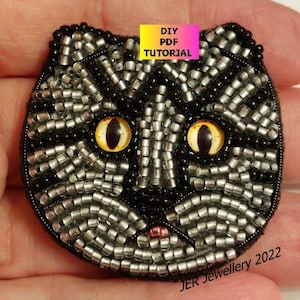 以下が含まれることがあります： 黒と銀のビーズでできた猫の顔のクローズアップ。大きな黄色の目があります。猫の顔は黒い背景にあり、猫の顔の上に「DIY PDF TUTORIAL」というテキストが黄色いボックスに入っています。「JER Jewellery 2022」というテキストは画像の下にあります。