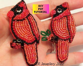 Puede incluir: Dos broches de pájaro cardenal hechos con cuentas de semillas rojas y negras. Los pájaros están posados sobre pequeñas ramas. Se ve una etiqueta rectangular amarilla y rosa con el texto DIY PDF TUTORIAL y ER Jewellery 2022.