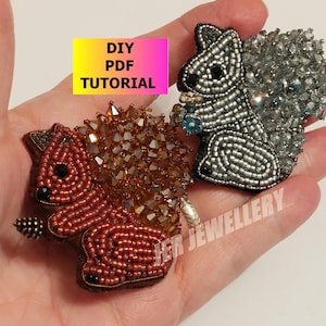 Może przedstawiać: Dwa haftowane broszki w kształcie zwierząt, jedna to brązowa wiewiórka z brązowym i złotym koralikowym ciałem, a druga to srebrna wiewiórka ze srebrnym i niebieskim koralikowym ciałem. Broszki znajdują się na dłoni, a w tle widnieje tekst "DIY PDF TUTORIAL".