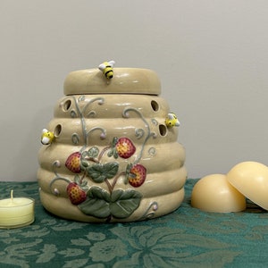 May include: A ceramic beehive-shaped wax warmer with a strawberry design and small bee figurines. The warmer is on a green tablecloth with two wax melts.