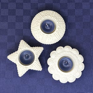 以下が含まれることがあります： ヒトデ、ウニ、花の形をした白いセラミック製のキャンドルホルダー3つ。各ホルダーには青いキャンドルが入っています。
