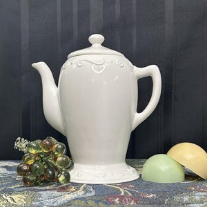 May include: White ceramic teapot with an ornate design and a wide base. The teapot is sitting on a patterned surface with green grapes and two round, yellow and green objects.