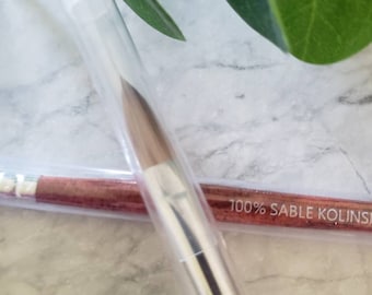 May include: A close-up of a makeup brush in a clear plastic tube. The brush is made of 100% sable kolinsky hair and has a silver ferrule. The tube has a red and gold label that says "100% Sable Kolinsky".