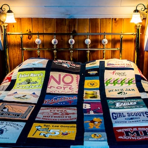 Peut inclure: Une courtepointe en patchwork faite de vieux t-shirts. La courtepointe est composée de différentes couleurs et motifs, notamment le bleu, le vert, le rouge et le jaune. La courtepointe est posée sur un lit avec une tête de lit en laiton. La courtepointe présente divers textes, notamment "Saint Mary's Carnival", "No More Excuses", "Phillies", "Beach Jam", "Girl Scouts", et "Myrtle Beach MB".