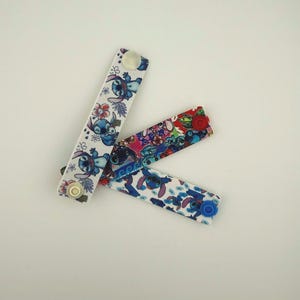Puede incluir: Tres extensores de botón de tela con patrones coloridos. Uno es blanco con personajes Stitch azules y flores, otro es una vibrante mezcla de colores, y el tercero es azul con Stitch. Cada uno tiene un cierre de botón.