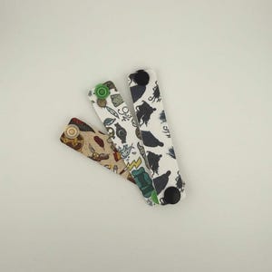 May include: Three patterned fabric wristbands. One is tan with gold details, another is white with black silhouettes, and the third is white with green and blue accents. Each has a button fastening.