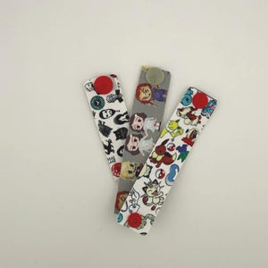 Puede incluir: Tres marcapáginas de tela con cierres de botón. Uno es blanco con imágenes en negro y rojo, otro es gris con imágenes de muñecas y el tercero es blanco con personajes de dibujos animados coloridos. Cada marcapáginas tiene un botón de diferente color.