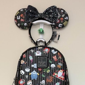 May include: A black backpack with a Nightmare Before Christmas theme, featuring character illustrations. A matching headband with a sequined bow and Mickey Mouse ears is displayed above. The headband has a green character charm.