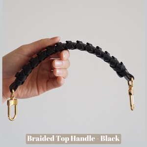 May include: A black braided leather top handle with gold metal clasps.  The handle is shown being held by a hand.