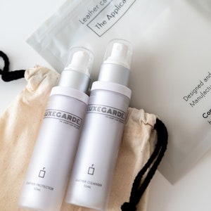 May include: Two white bottles with silver caps and labels that say "Luxegarde" and "The Luxury Car Specialist". The bottles are labelled "Leather Protector 50ml" and "Leather Cleanser 50ml". The bottles are in a white drawstring bag with black drawstrings. A white bag with the text "Leather care cloth The Applicator" is in the background.
