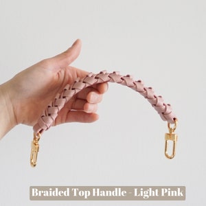 May include: A light pink braided top handle with gold metal clasps. The handle is made of woven leather and is designed to be used on a handbag or purse.