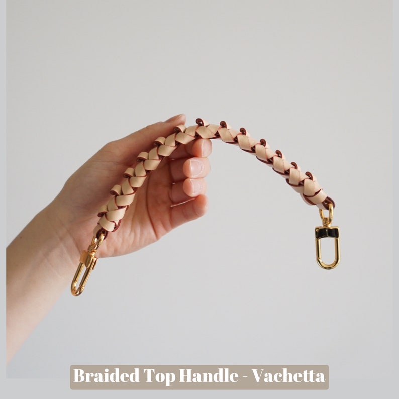 May include: A braided leather top handle in a light beige and burgundy color. The handle has a gold metal clasp.