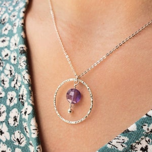 Puede incluir: Un collar de plata con un colgante circular texturizado y una piedra preciosa p&uacute;rpura que cuelga de la parte inferior.