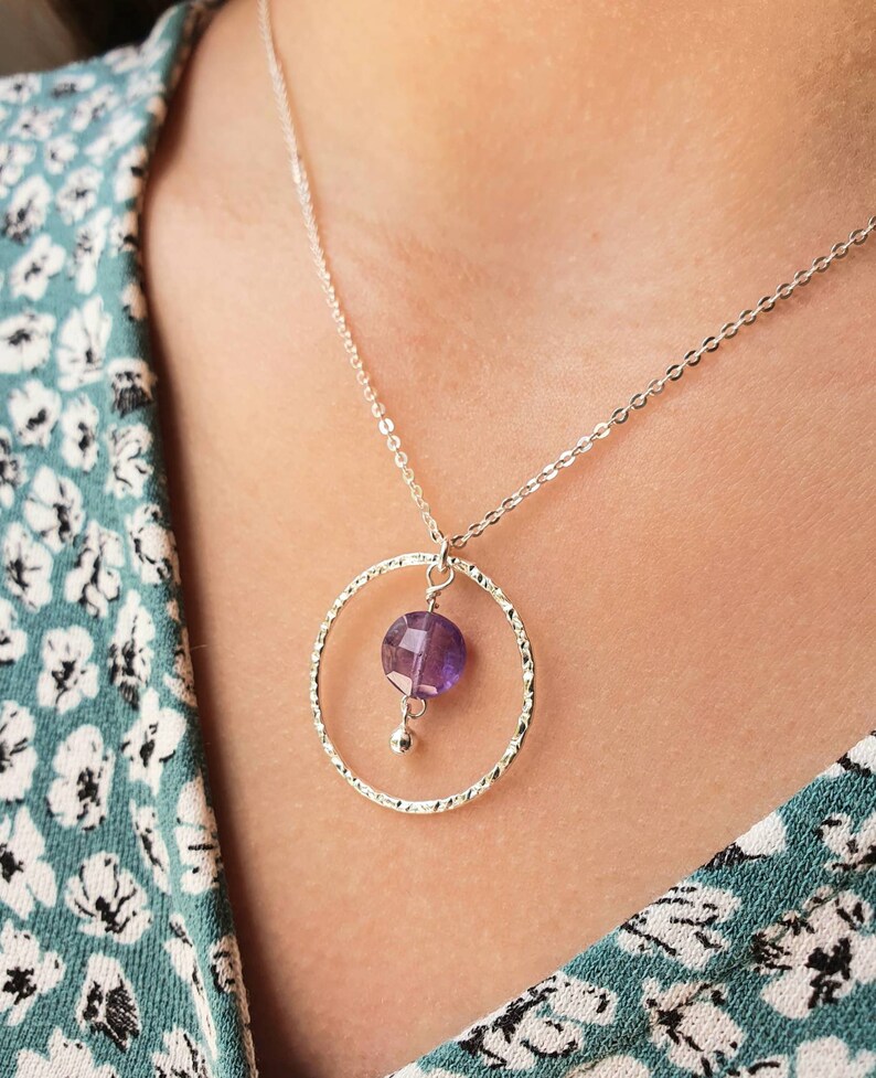 Puede incluir: Un collar de plata con un colgante circular texturizado y una piedra preciosa p&uacute;rpura que cuelga de la parte inferior.