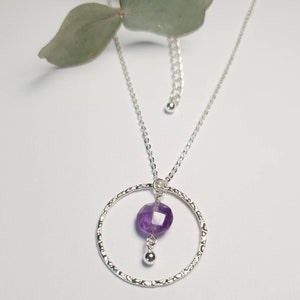 Puede incluir: Un collar de plata con un colgante circular texturizado y una piedra preciosa de amatista p&uacute;rpura que cuelga de la parte inferior.