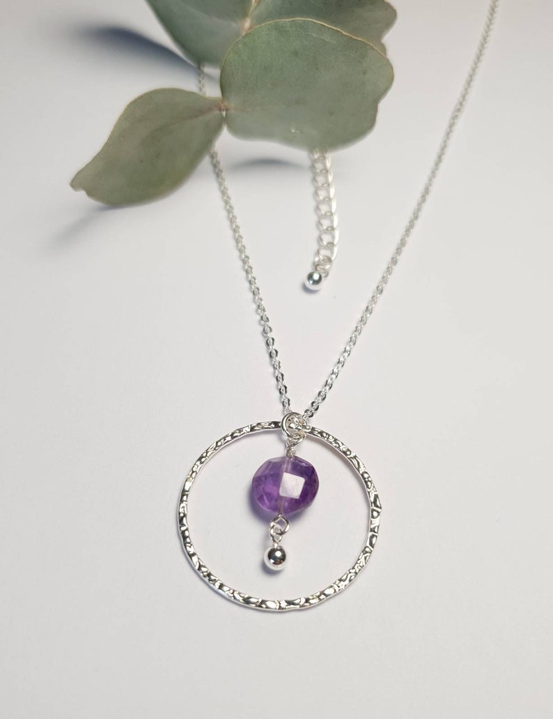 Puede incluir: Un collar de plata con un colgante circular texturizado y una piedra preciosa de amatista p&uacute;rpura que cuelga de la parte inferior.