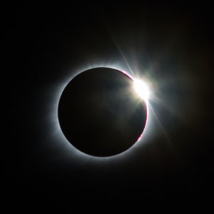 May include: A photograph of a solar eclipse, showing the moon covering the sun. A bright corona surrounds the dark disc of the moon, with a burst of light on the right. The background is a deep black, creating a dramatic contrast in this celestial event.