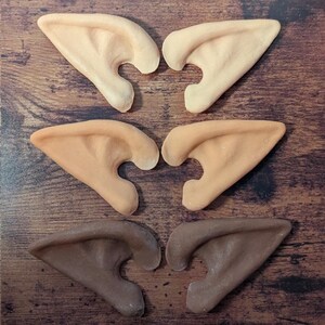 Puede incluir: Seis pares de orejas de elfo en diferentes tonos de piel, dispuestas sobre una superficie de madera. Las orejas varían en color desde beige claro hasta marrón medio, mostrando una gama de tonos de piel. Accesorios de disfraz para cosplay o eventos temáticos.