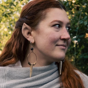 May include: A woman with long red hair and elf ears is wearing gold hoop earrings. The earrings feature a dark bead at the top, a gold hoop, and a light-colored antler pendant. She is wearing a gray sweater.