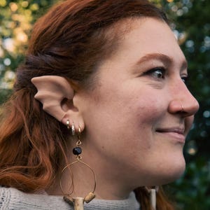 May include: A side profile of a person wearing a pair of pointed elf ear prosthetics and a gold hoop earring with a black bead and a bone-like pendant. The person has red hair and is wearing a gray sweater.
