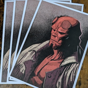 May include: Several art prints showcasing a comic book illustration of a red-skinned humanoid character with horns. The character is wearing a dark coat and has a serious expression. The prints have white borders and are stacked on a wooden surface.