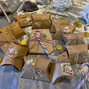 Op de afbeelding: Een feestelijke opstelling van kleine, bruine geschenkdozen, vastgebonden met touw en versierd met kerststickers. De cadeaus zijn gerangschikt rond een groter, verpakt cadeau, zuurstokken en gouden munten.
