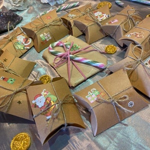 Op de afbeelding: Een verzameling kleine, bruine geschenkdozen vastgebonden met touw, versierd met kerststickers en gerangschikt op een glinsterend oppervlak. Gouden chocolademunten en zuurstokken voegen feestelijke accenten toe. Witte glitterrozen en een donkerpaarse kristal op de achtergrond.