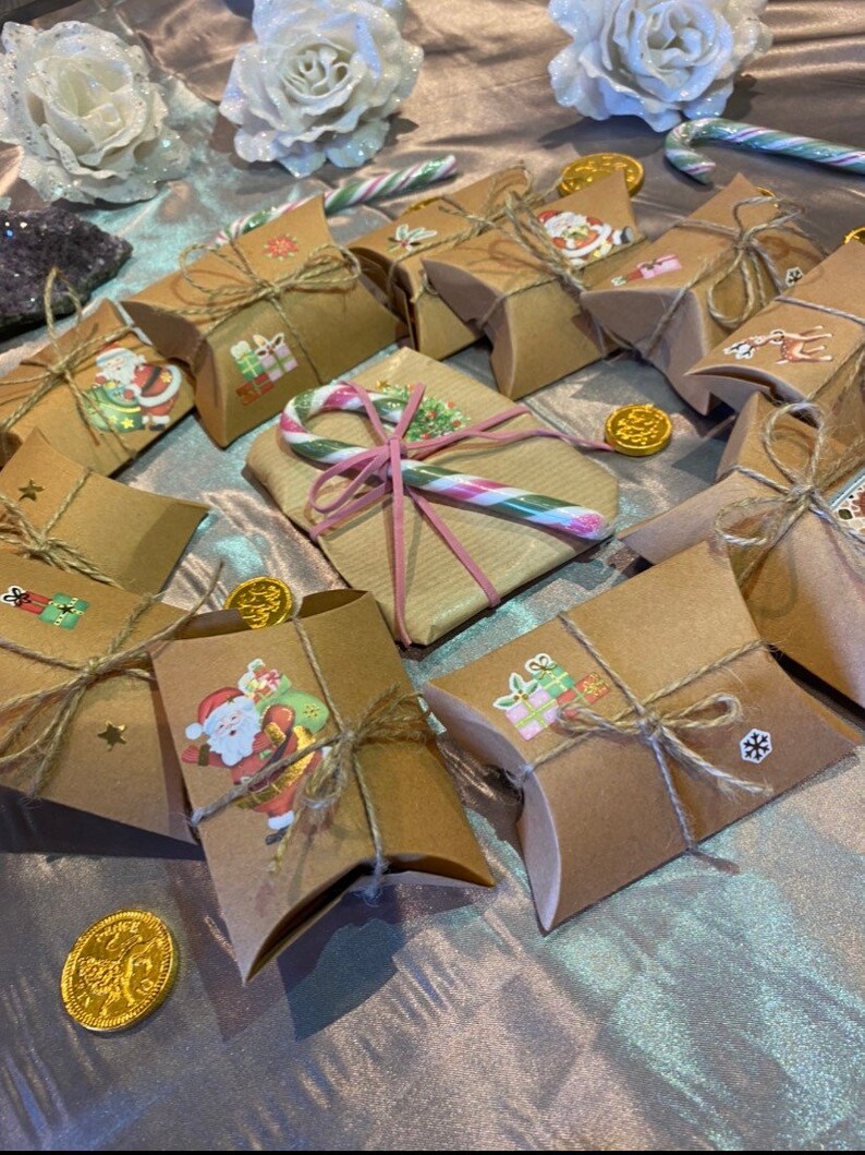 Op de afbeelding: Een verzameling kleine, bruine geschenkdozen vastgebonden met touw, versierd met kerststickers en gerangschikt op een glinsterend oppervlak. Gouden chocolademunten en zuurstokken voegen feestelijke accenten toe. Witte glitterrozen en een donkerpaarse kristal op de achtergrond.