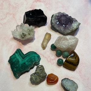 Op de afbeelding: Een assortiment van verschillende kristallen en edelstenen, waaronder amethist, kwarts, malachiet en obsidiaan. De kristallen hebben verschillende vormen en maten, met kleuren vari&euml;rend van diep paars tot groen en helder. Er zijn ook verschillende kleine, groene, ronde stenen.