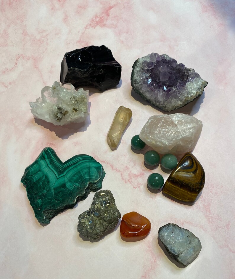 Op de afbeelding: Een assortiment van verschillende kristallen en edelstenen, waaronder amethist, kwarts, malachiet en obsidiaan. De kristallen hebben verschillende vormen en maten, met kleuren vari&euml;rend van diep paars tot groen en helder. Er zijn ook verschillende kleine, groene, ronde stenen.