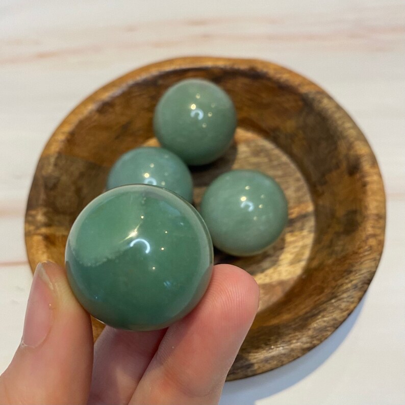 Op de afbeelding: Vier groene bollen met een gepolijst oppervlak in een houten kom. De bollen vari&euml;ren enigszins in tint, met &eacute;&eacute;n in een hand. De kom heeft een natuurlijke houtnerf. De bollen zijn ongeveer 3-4 cm in diameter.